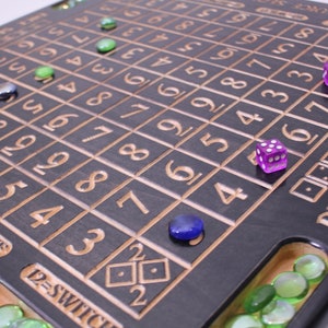 May include: A wooden Dice Sequence board game with a dark finish and engraved numbers. The board has a grid pattern with numbers and text. Several colored glass marbles and two purple dice are on the board. The game is designed for multiple players.