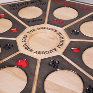 May include: An octagonal wooden game board featuring a light natural wood center engraved with "THE BULLEE'S RUMMOLT AUGUST 23RD, 2025". The board has dark grey sections with circular cutouts, red and black card suit symbols, and text including "SEQUENCE", "POKER POT", and "KING".