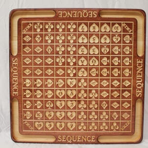 May include: A wooden game board with a brown and light wood finish. The board is marked with a grid of squares, each containing a playing card symbol. The board is labeled "SEQUENCE" on all four sides.