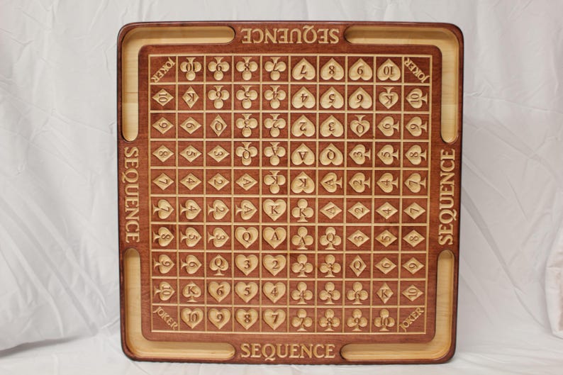 May include: A wooden game board with a brown and light wood finish. The board is marked with a grid of squares, each containing a playing card symbol. The board is labeled "SEQUENCE" on all four sides.