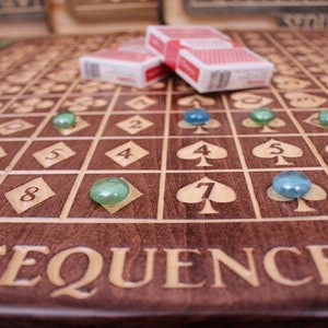 May include: A wooden game board with the word "SEQUENCE" engraved on it. The board has a grid of squares with numbers and playing cards symbols. There are green and blue glass marbles on the board.
