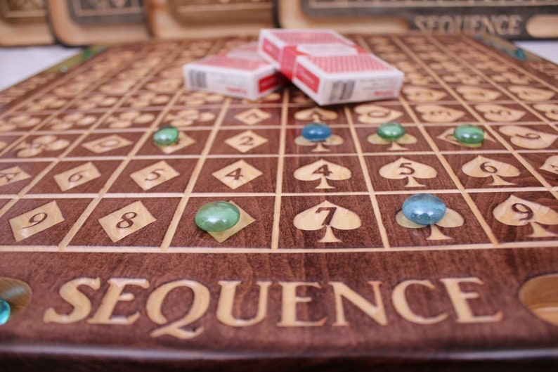 May include: A wooden game board with the word "SEQUENCE" engraved on it. The board has a grid of squares with numbers and playing cards symbols. There are green and blue glass marbles on the board.