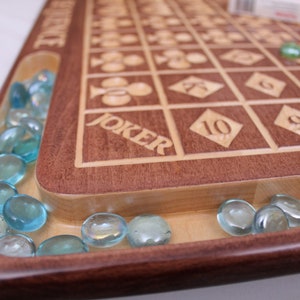 Pu&ograve; includere: Un tabellone da gioco in legno con una griglia di quadrati, ognuno contenente un numero o un simbolo di carta da gioco. Il tabellone ha una scanalatura attorno al bordo riempita di biglie di vetro blu. La parola "JOKER" &egrave; incisa sul tabellone.