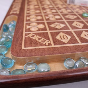 May include: A wooden game board with a light brown surface and a dark brown border. The board has a grid of squares with numbers and symbols engraved on them. The word "JOKER" is engraved on the board. The board has a groove around the edge filled with blue glass marbles.