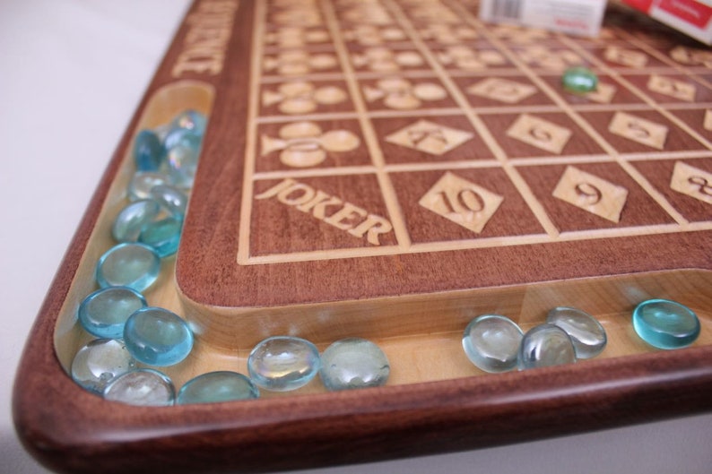 May include: A wooden game board with a light brown surface and a dark brown border. The board has a grid of squares with numbers and symbols engraved on them. The word "JOKER" is engraved on the board. The board has a groove around the edge filled with blue glass marbles.