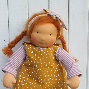 Könnte beinhalten: Eine handgefertigte Puppe mit roten Haaren, die ein gelbes Kleid mit weißen Punkten, ein rosa-weiß gestreiftes Hemd und braune gehäkelte Schuhe trägt. Die Puppe steht auf einem Holzsockel.
