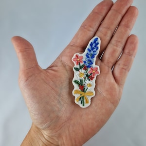 Może przedstawiać: Naszywkę z białego filcu z haftowanym bukietem kwiatów. Bukiet zawiera niebieskie, różowe i białe kwiaty z zielonymi liśćmi i żółtą wstążką.