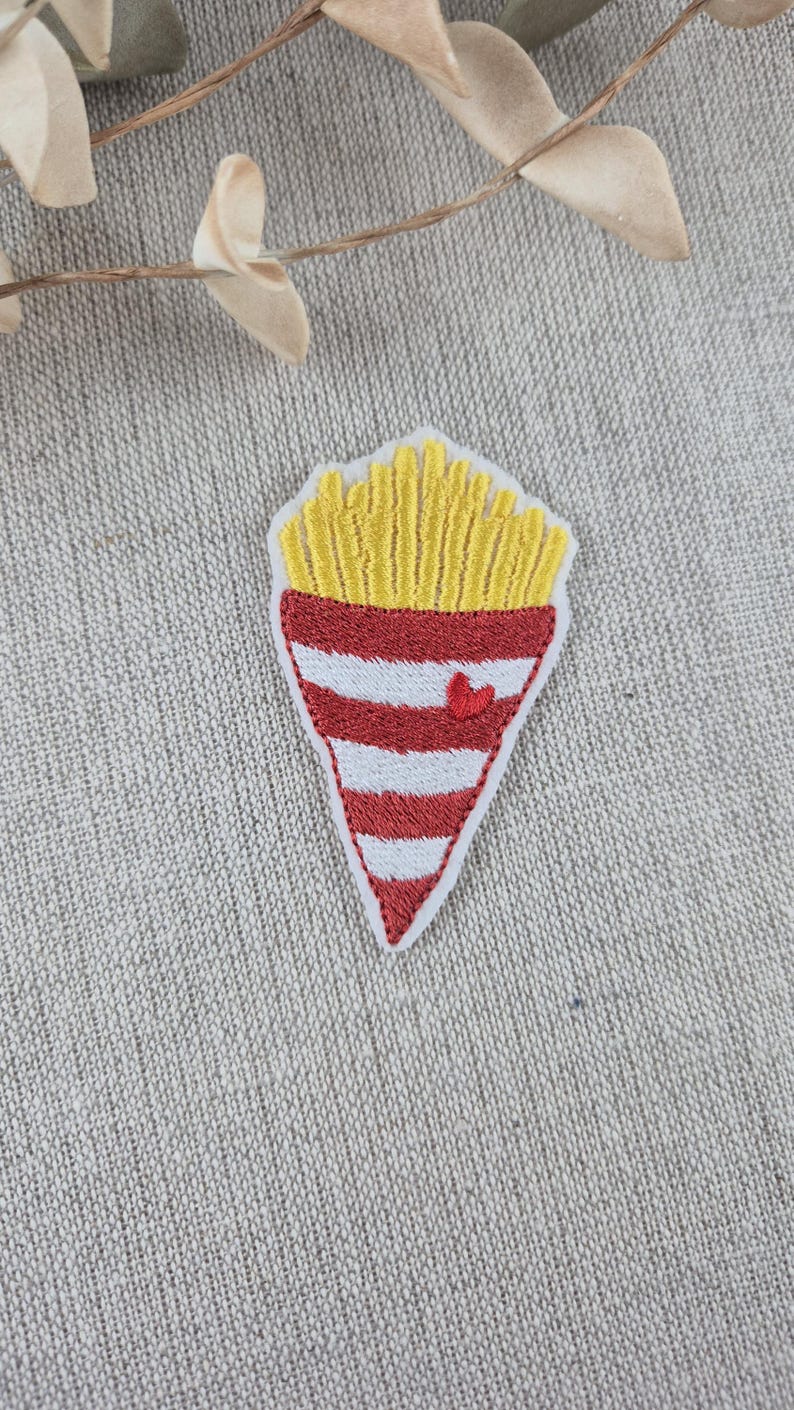 May include: Embroidered patch featuring a cone of golden french fries. The cone is red and white striped with a small red heart. The patch is on a neutral fabric background.