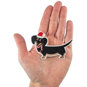 Può includere: Un bassotto nero e marrone con un cappello di Babbo Natale rosso e una sciarpa rossa e bianca. Il bassotto è ricamato su una toppa di tessuto bianco.