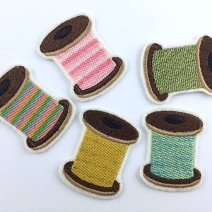 Pode incluir: Cinco patches bordados para passar a ferro com forma de rolos de fio. Os rolos são de várias cores e padrões, incluindo riscas rosa e brancas, riscas verdes e laranja, amarelo e azul e verde.