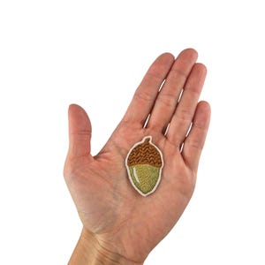 Może przedstawiać: Naszywka z haftowanej żołędzi w kolorze brązowym i zielonym. Naszywka znajduje się na dłoni, ale skupiamy się na naszywce.