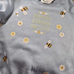 May include: A light blue sweatshirt with embroidered bees and flowers. The text "Bienchen & Bl&uuml;mchen und sssssso" is embroidered on the front.