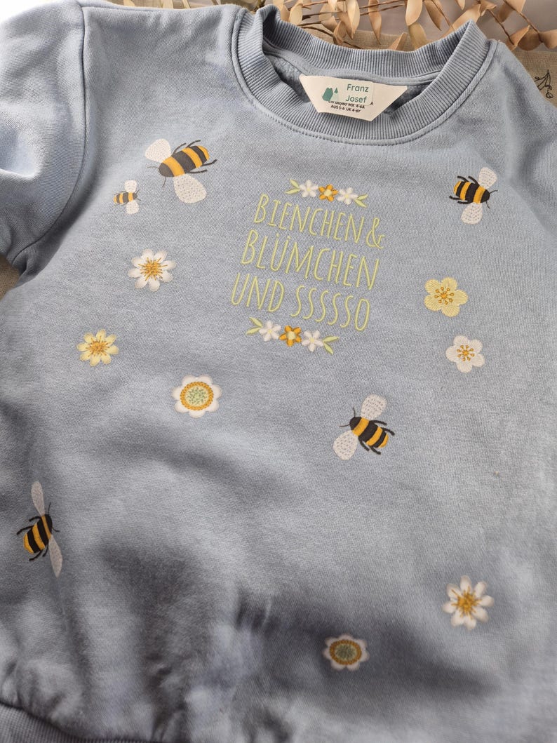 May include: A light blue sweatshirt with embroidered bees and flowers. The text "Bienchen & Bl&uuml;mchen und sssssso" is embroidered on the front.
