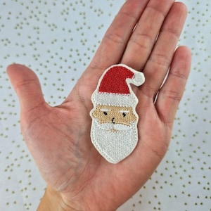 Könnte beinhalten: Ein weiß gestickter Weihnachtsmann-Patch mit roter Mütze und weißem Bart. Der Patch befindet sich auf einer Hand.