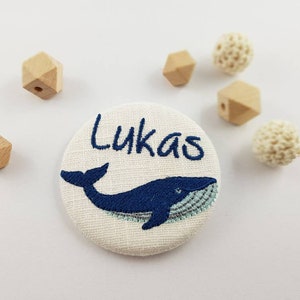 Peut inclure: Un bouton en tissu blanc avec une broderie bleue d'une baleine et le nom "Lukas".