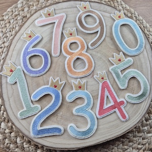 Può includere: Dieci toppe di feltro colorate con numeri ricamati da 1 a 10. Ogni numero ha una corona dorata con un cuore rosso in cima. Le toppe sono disposte in cerchio su una superficie di legno.