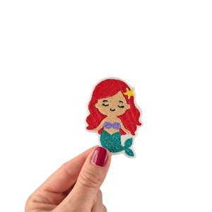 May include: Embroidered patch of a cartoon mermaid with red hair, a purple top, and a teal tail. A yellow star is in her hair. The patch is held up by a hand against a white background.