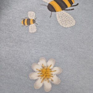 May include: A light blue fabric with a white daisy and two embroidered bees. The larger bee is yellow and black with white wings. The smaller bee is yellow and black with white wings.