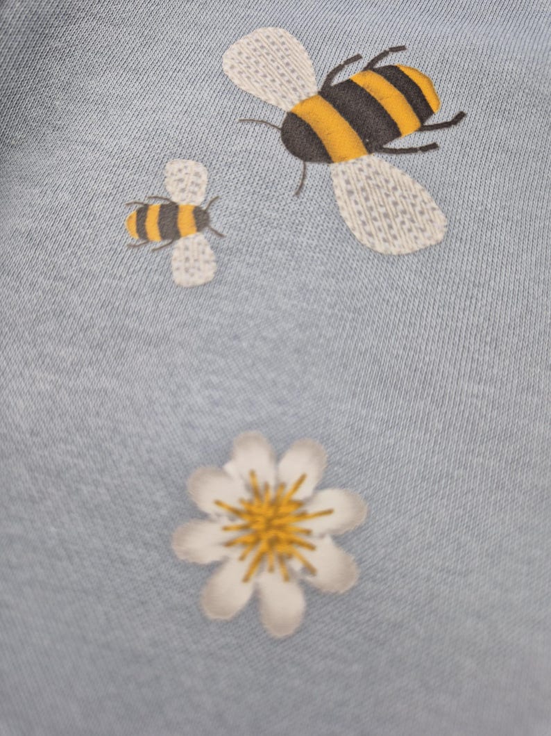 May include: A light blue fabric with a white daisy and two embroidered bees. The larger bee is yellow and black with white wings. The smaller bee is yellow and black with white wings.