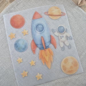 Könnte beinhalten: Aquarell-Illustration im Weltraum-Design mit einer blau-roten Rakete, Planeten, Sternen und einem Astronauten. Das Design hat eine weiche Pastell-Farbpalette mit Orange-, Gelb- und Blautönen. Die Elemente sind auf einem durchscheinenden Hintergrund angeordnet.