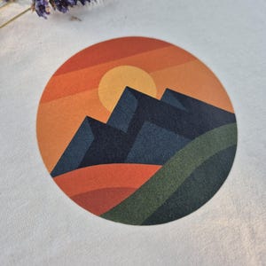 Könnte beinhalten: Cremefarbenes T-Shirt mit einem kreisförmigen Berglandschaftsmotiv. Das Design zeigt eine gelbe Sonne, einen orange-roten Himmel und dunkelblaue Berge. Grüne und orangefarbene Hügel vervollständigen die Szene. Getrocknete Lavendelzweige befinden sich oben links.