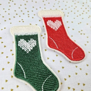 Op de afbeelding: Twee geborduurde kerstsok-patches, een groene en een rode, met witte viltbies en een wit hartontwerp.
