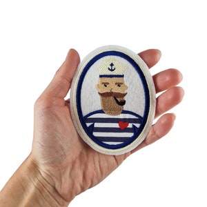 May include: Oval embroidered patch featuring a cartoon sailor with a beard, pipe, and anchor on his hat. The sailor wears a blue and white striped shirt and has a red heart. The patch is held in a hand against a white background.