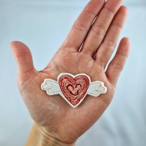 May include: A red heart-shaped patch with embroidered details and small white wings. The heart has a smaller heart design in the center. The patch is held in an open hand against a white background.