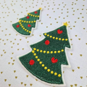 Könnte beinhalten: Zwei grüne Filz-Weihnachtsbaum-Patches mit roten und gelben Stickereien. Die Bäume befinden sich auf einem weißen Hintergrund mit einem goldenen Dreiecksmuster.