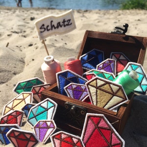 May include: A wooden treasure chest filled with colorful embroidered diamond patches. The chest is open and the patches are spilling out. The chest is sitting on a sandy beach with a sign that says "Schatz" in the background.