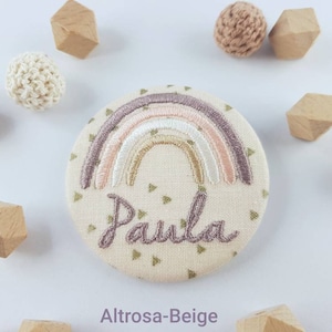 Op de afbeelding: Een beige stoffen pin met een regenboogontwerp en de naam "Paula" geborduurd in paarse draad. De pin is omringd door kleine houten en gehaakte decoraties.