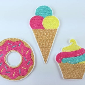 Könnte beinhalten: Drei gestickte Aufnäher mit einem rosafarbenen und weißen Donut, einem gelben, rosafarbenen und blauen Eiswaffel und einem gelben, rosafarbenen und blauen Cupcake.