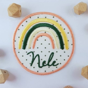 Könnte beinhalten: Ein weißer Stoff-Patch mit einem Regenbogen-Design, das in Gelb, Grün und Orange gestickt ist. Der Patch hat einen weißen Hintergrund mit schwarzen Punkten und den Namen "Nele" in Grün gestickt.