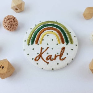 Könnte beinhalten: Ein weißer Stoffkreis mit einem grünen, braunen und gelben gestickten Regenbogen und dem Namen Karl in schwarzer Stickerei.