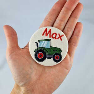 Könnte beinhalten: Runder, gestickter Aufnäher mit einem grünen Traktor, schwarzen Rädern und roten Akzenten. Der Name "Max" ist rot über dem Traktor gestickt. Der Aufnäher besteht aus beigem Stoff.