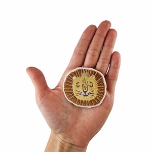 Könnte beinhalten: Ein gestickter Löwenkopf-Patch in Braun und Gold mit weißem Rand.