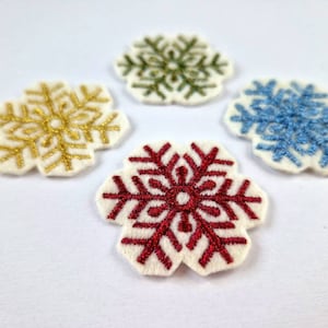 Może przedstawiać: Cztery haftowane naszywki w kształcie płatków śniegu w kolorze złotym, zielonym, niebieskim i czerwonym. Każdy płatek śniegu jest wyszyty na białej filcowej podstawie. Naszywki ułożone na białej powierzchni, idealne do zimowych dekoracji.