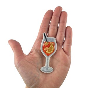 Könnte beinhalten: Ein gestickter Bügel-Patch mit einem orangenen Getränk in einem Glas mit Strohhalm und Orangenscheibe. Der Patch ist weiß mit orangener, gelber und brauner Stickerei.