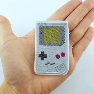 Può includere: Un ricamo a forma di console per videogiochi portatile. La console è di colore bianco e grigio, con uno schermo grigio con un cuore rosa nell'angolo in alto a sinistra e uno schermo giallo al centro. La console ha un pulsante più grigio e due pulsanti circolari rosa.