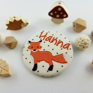 Op de afbeelding: Een witte stoffen knoop met een bruine vos erop geborduurd. De knoop heeft witte stippen en de naam "Hanna" is geborduurd in zwarte draad.