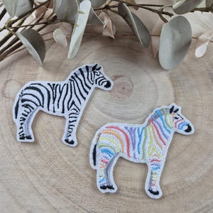 Può includere: Due toppe ricamate da stirare a forma di zebra. Una zebra è bianca e nera, l'altra è una zebra arcobaleno.