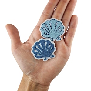 Op de afbeelding: Twee geborduurde strijkpatches in de vorm van schelpen. De grotere schelp is lichtblauw met een witte omtrek. De kleinere schelp is donkerblauw met een witte omtrek.