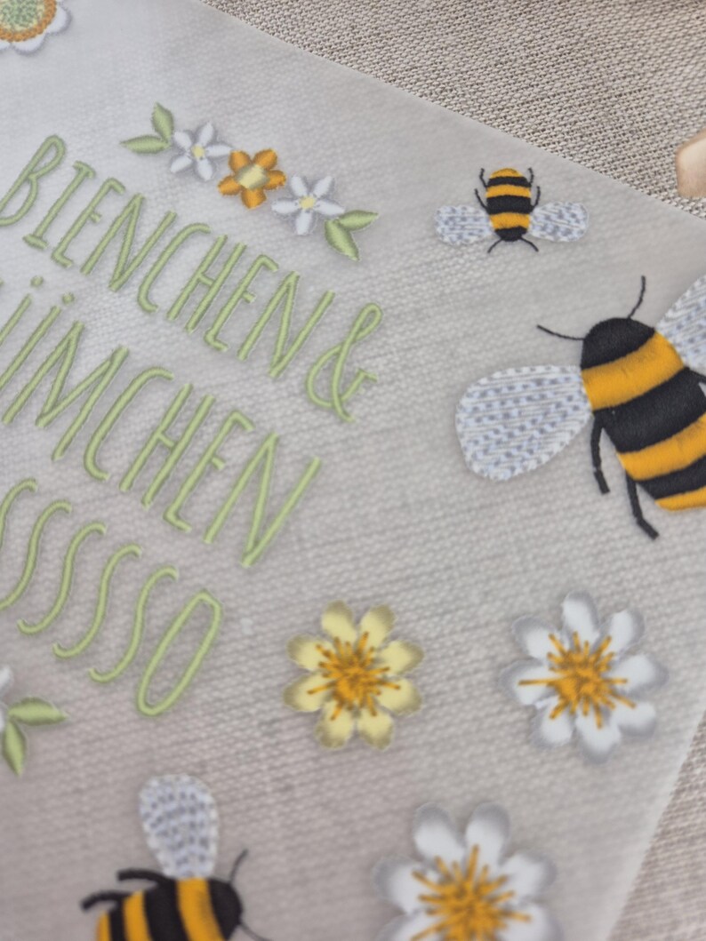 May include: A white card with the text "Bienchen & Imchen sssssso" in green embroidery. The card is decorated with yellow and white flowers and bees.
