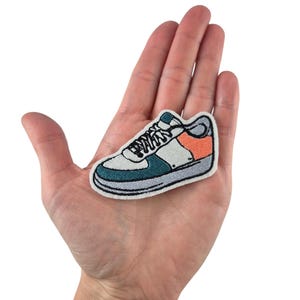 Peut inclure: Écusson brodé d'une basket blanche, turquoise, orange et grise. Le patch est une version miniature d'une chaussure de sport classique, avec des lacets et un design détaillé. Le patch est tenu dans une main.