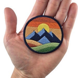 Könnte beinhalten: Gestickter Aufnäher mit einer Berglandschaft bei Sonnenuntergang. Das Design zeigt blaue Berge, eine gelbe Sonne und einen Himmel mit Farbverlauf in Orange- und Brauntönen. Der Aufnäher ist rund mit einem marineblauen Rand.