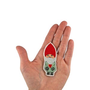 May include: A red and green embroidered iron-on patch of a gnome with a red hat, white beard, and a red heart on its chest.