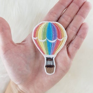 Könnte beinhalten: Ein gestickter Aufnäher mit einem Regenbogen-Heißluftballon. Der Ballon ist mit einem Regenbogen aus Farben gefüllt, darunter Rot, Orange, Gelb, Grün, Blau, Indigo und Violett. Der Ballon hat einen weißen Korb mit schwarzen Linien.