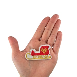 Könnte beinhalten: Ein gestickter Weihnachts-Schlitten-Patch in Rot und Weiß mit einem goldenen Herzen und goldenen Verzierungen. Der Patch wird in der Hand einer Person gehalten.