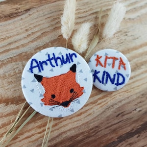 Könnte beinhalten: Zwei gestickte Stoff-Patches. Ein Patch zeigt ein Fuchses-Gesicht mit dem Namen "Arthur" darüber. Der andere Patch sagt "Kita Kind".