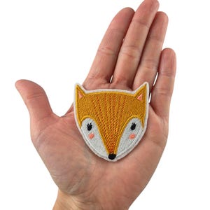 Könnte beinhalten: Ein gelber gestickter Fuchskopf-Patch mit weißen Akzenten und rosa Wangen.